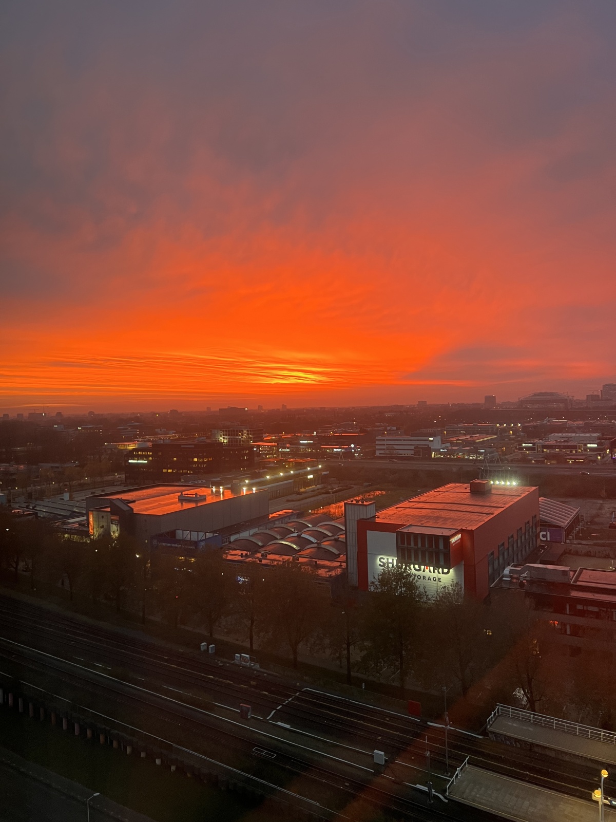 A breathtaking sunrise over Overamstel with a rich gradient of red and orange filling the sky above the cityscape and railway tracks, as seen from the Leonardo Hotel room. The canal is not visible in the shot.