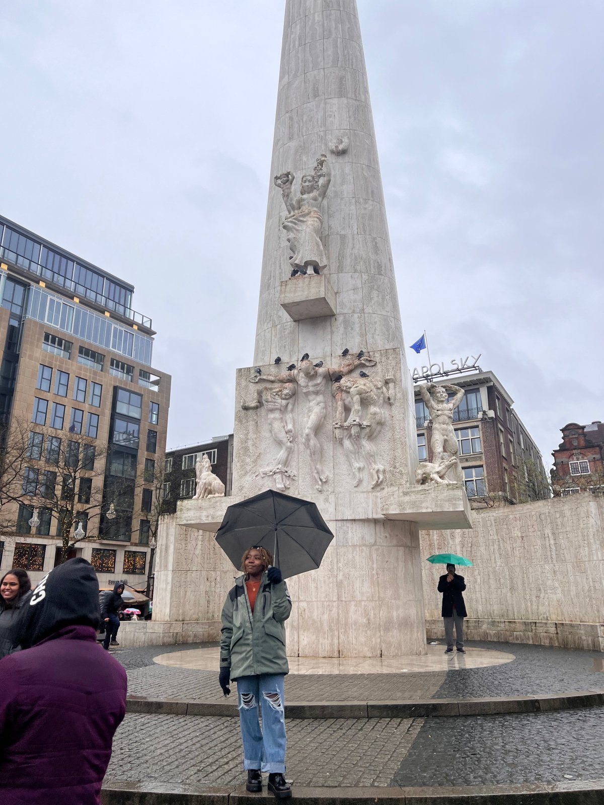 Person standing with an umbrella in front of the National Monument on Dam Square in Amsterdam, with the Royal Palace visible in the background.